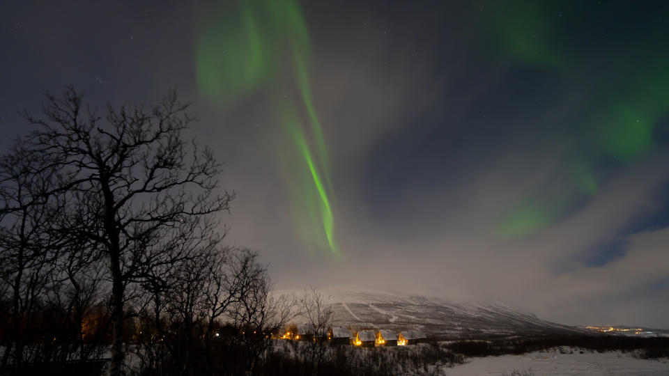 bands of green light in a partially clouded sky above small huts with lights on.