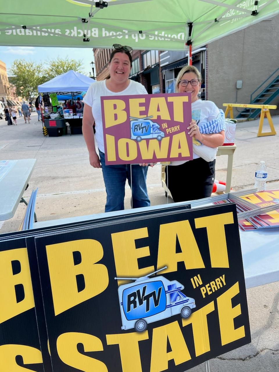 Andrea Brownlee and Martha Stetzel hold up a yard sign for sale at the Perry Farmers Market to help promote the RVTV Perry Tailgate, which will be held on Monday, Sept. 2 in downtown Perry.