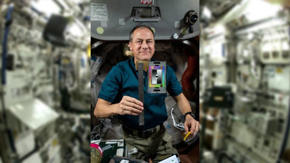  An inset photo of an astronaut with a square-shaped device showing colors and rulers. in back is a generic, blurred photo of the international space station showing a module with lots of items in it. 