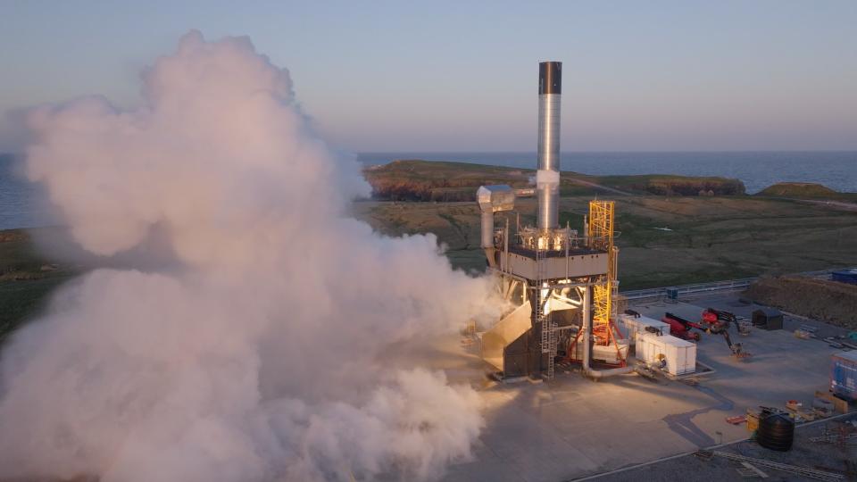  Fire and smoke spew from a sliver cylinder upright on a large metal platform. 