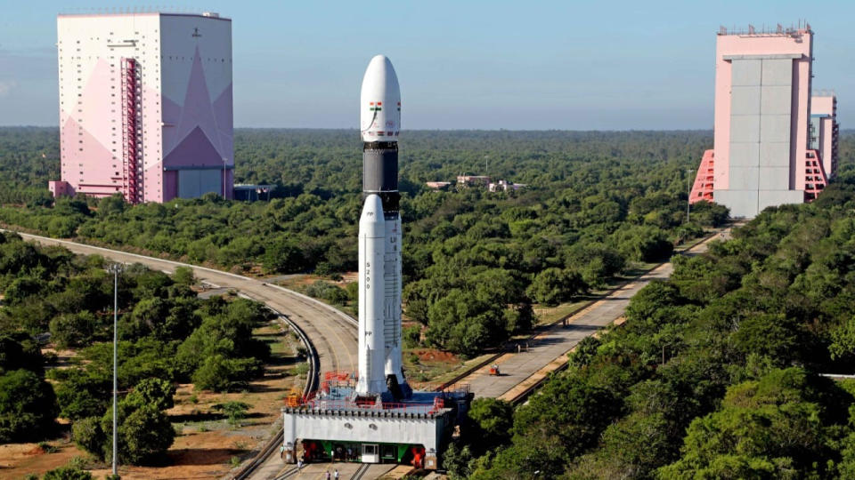  A white rocket with two white side boosters stands on a grey metal platform fixed on a track at the splitting toward two pink decorated hanger buildings. 