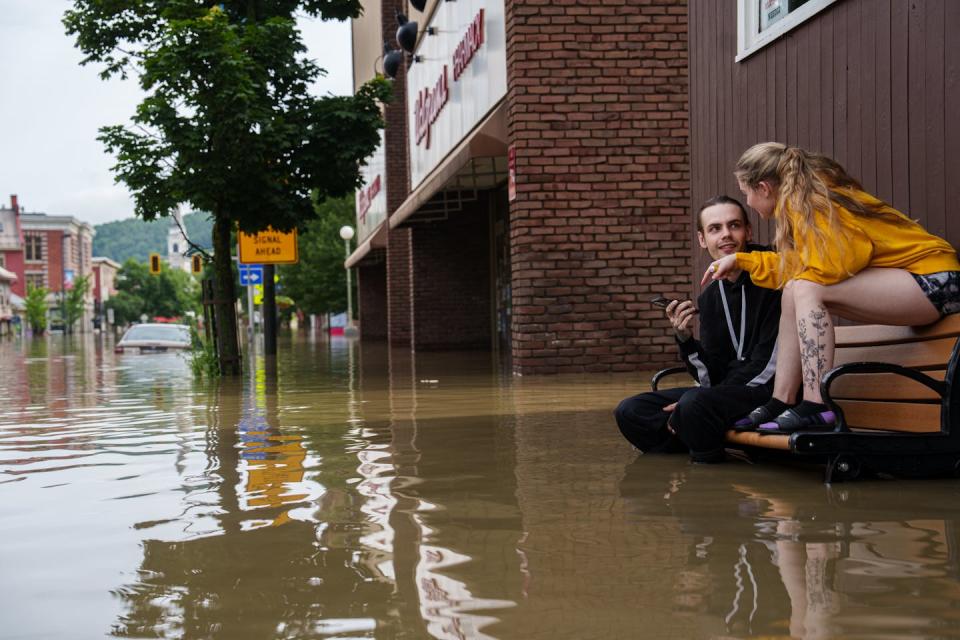 A man and woman sit on a park bench with water up to the  man's knees. The woman is sitting on the chair back. A car in the street is flooded up to the roof.