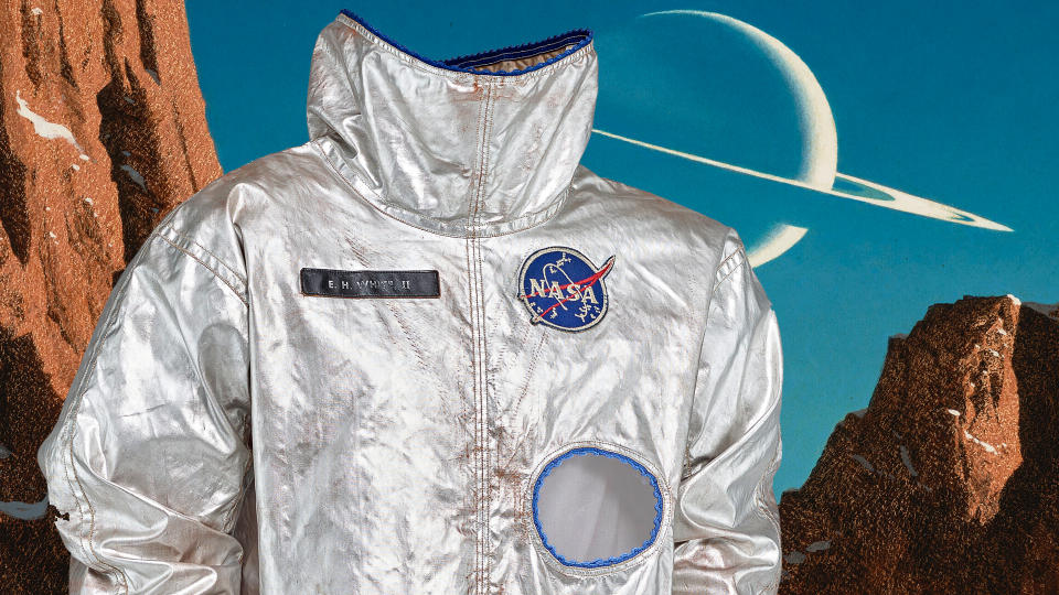  A silver flight suit without a helmet is seen against a backdrop of red rocks in front of a blue sky, with a large ringed planet hanging large in it. 