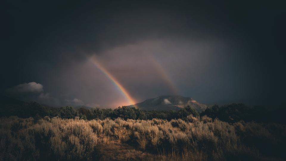  A faint but colorful double rainbow rises out from the landscape into a cloudy sky. . 