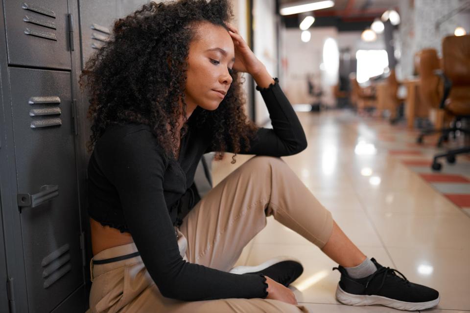 Female student sits in a school hallway with her back against a locker, looking forlorn.