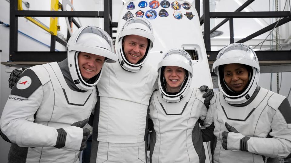  Four people in white spacesuits with the visors up pose and smile in front of a white cone-shaped spacecraft. 