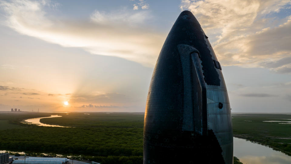  Closeup of a conical rocket nose towering above a wetland landscape at sunset. 