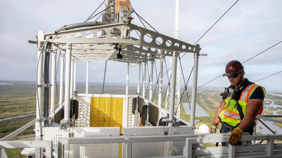  A large basket with a person beside in a reflective vest. behind is a landscape indicating they are quite a distance above the ground. 