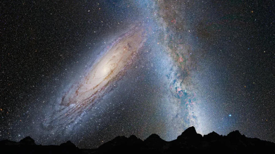  A silhouette of a mountainous horizon lays low on the image below a vast sky of stars with the arm of the milky way shooting up from the right, and a giant galaxy filling the left of the sky. 