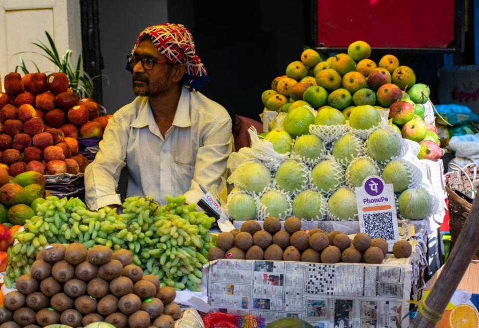 A man behind piles of fruits at his stall in India that features a QR payment code.