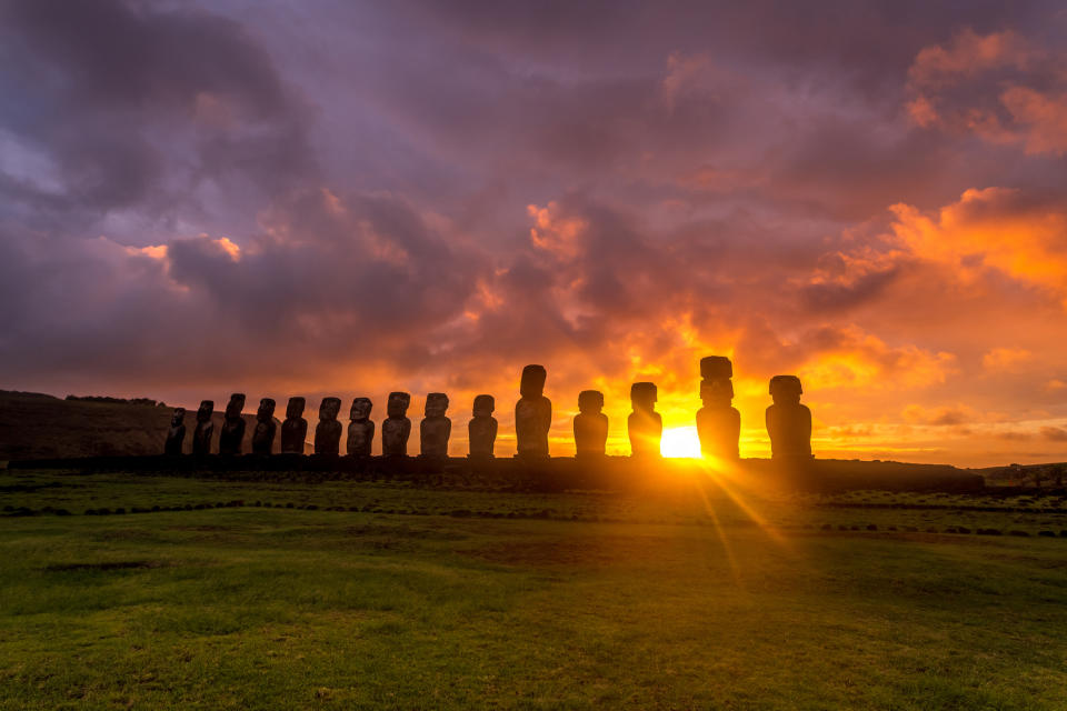 several dozen tall stone head statues stand on a lush green island under a colorful sunset