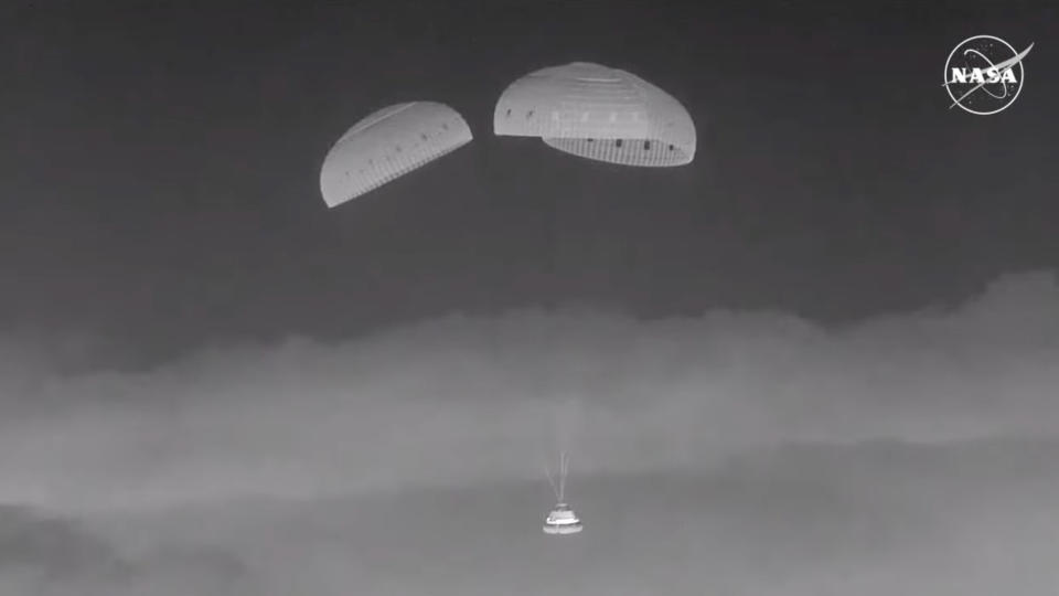  Black and white photo of a space capsule descending through the sky under three parachutes. 