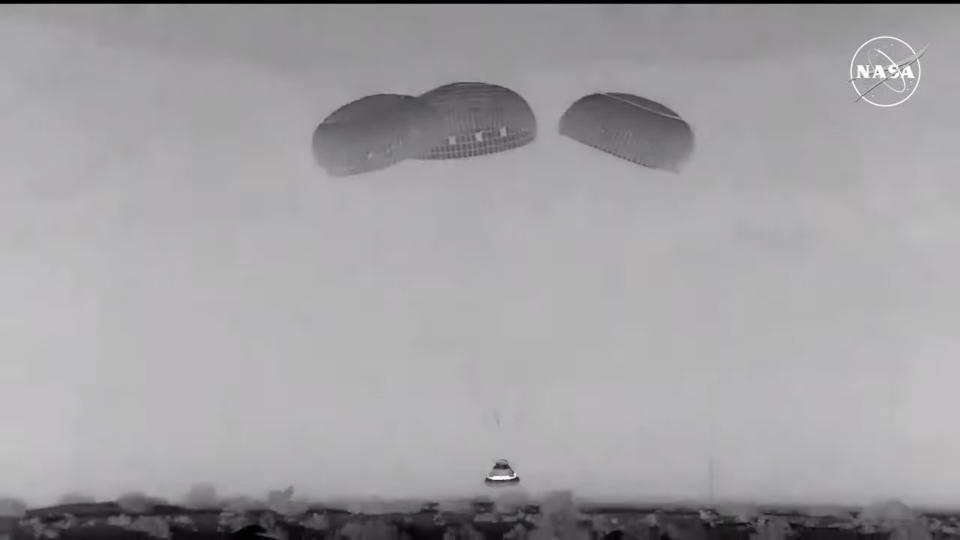 a black and white view of a capsule descending under parachutes. a nasa logo is at upper right as the broadcast ran on nasa television