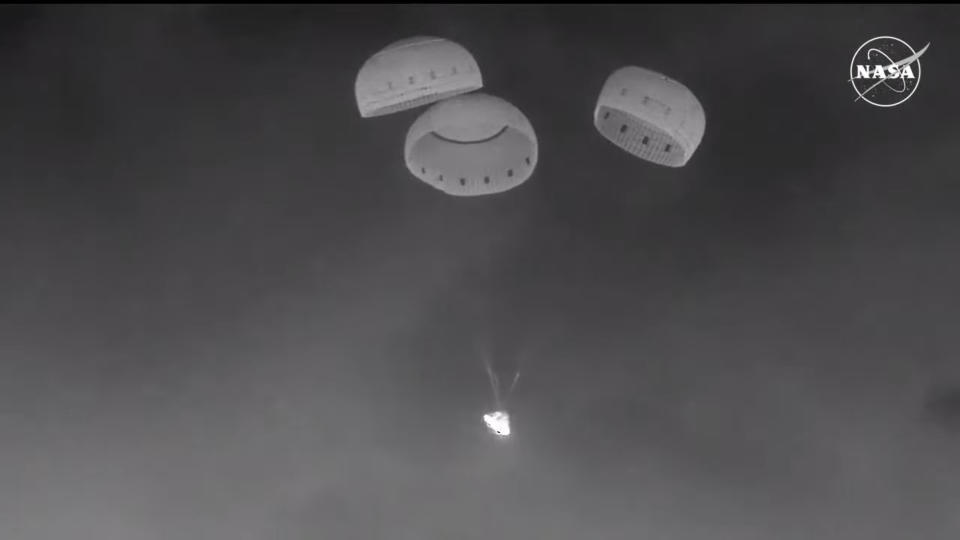  A black and white image of a cone-shaped spacecraft dangling under parachutes at night. 