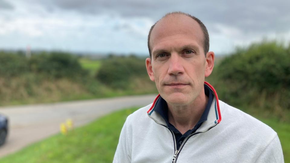 A man stands by a roadside, with green fields, a road and hedges in the background. 