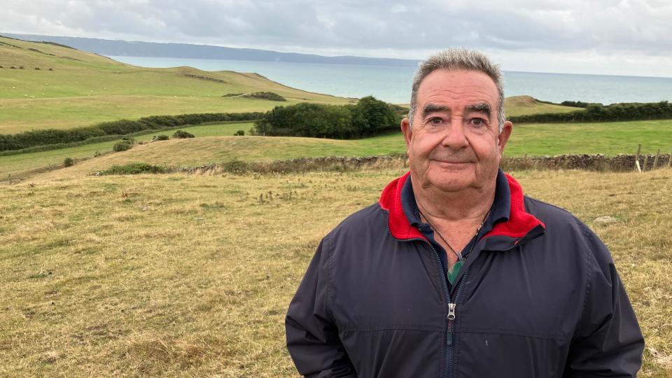 A farmer in a blue coat with red collar stands in a field close to the coast.  Rolling fields are in the background and beyond that the sea. 