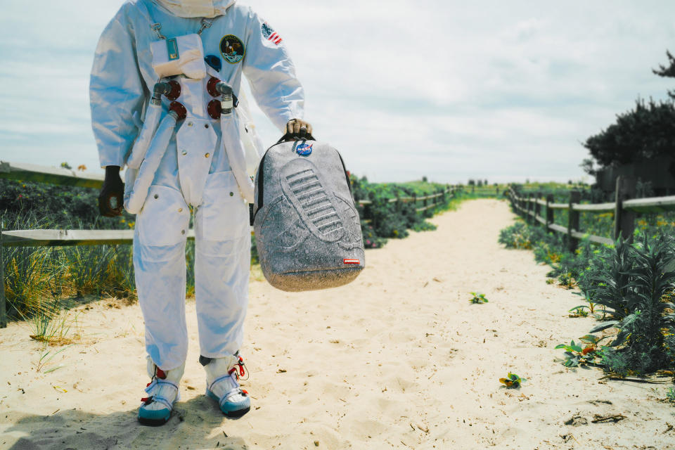 a model in a mock spacesuit walks on a beach carrying a backpack featuring a mottled grey look similar to the surface of the moon