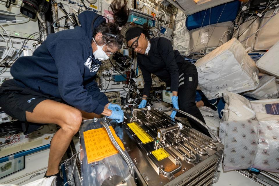 two women in sweat suits unpack a large metal box in a cramped laboratory full of wires and computers. they are floating