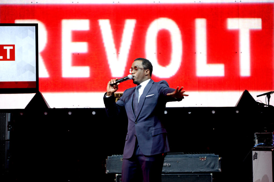 Combs speaks into a microphone in front of a backdrop with the Revolt logo.