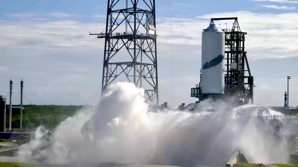  Outside on a sunny day, with some flat, white cloud cover, A small cylindrical tank with a large black feather painted on the side billows water and steam in a rush below. It is supported by a short metal structure, adjacent to a tall lighting tower. 