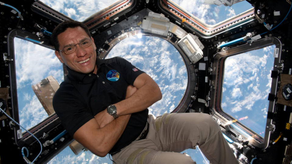 A man in khakis crosses his arms as he floats in front of a hexagonal window that looks down at Earth from orbit.