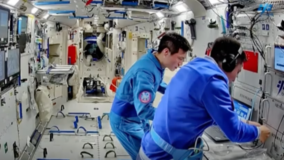  Two astronauts in blue flight suits look at a laptop aboard a white-walled space station. 