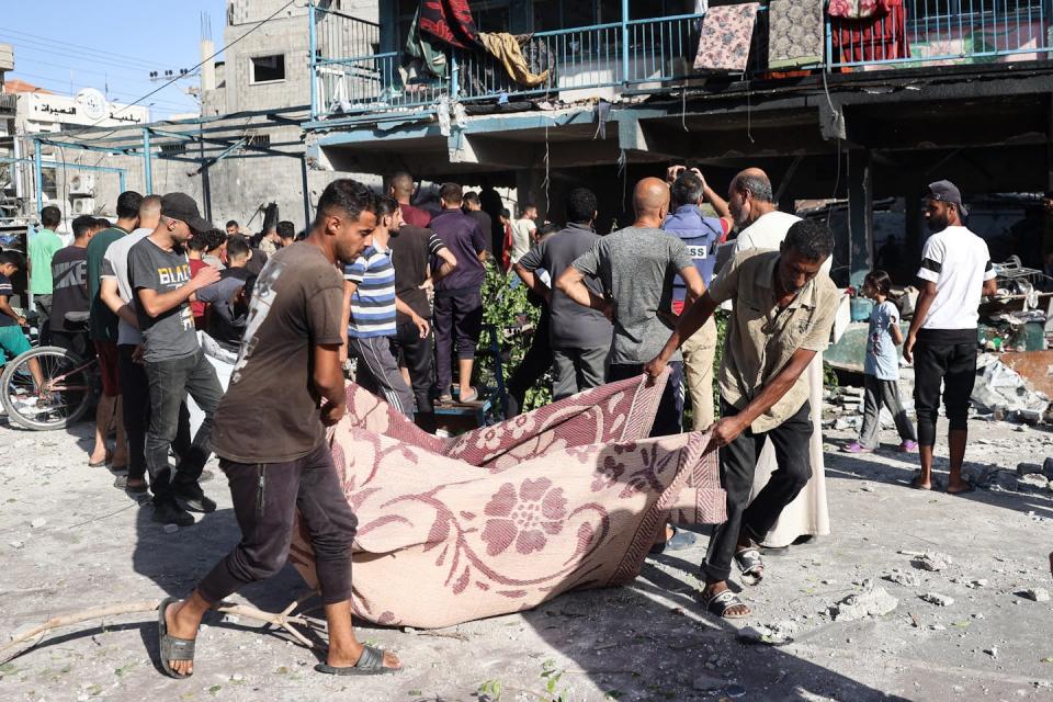 Two young men hold a blanket with a bombing victim inside as several dozen other people stand around damaged site of Israeli air strike.