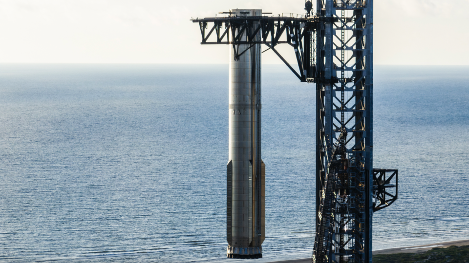  Closeup of a silver rocket booster held above ground by the mechanical arms of its launch tower, with the ocean in the background. 