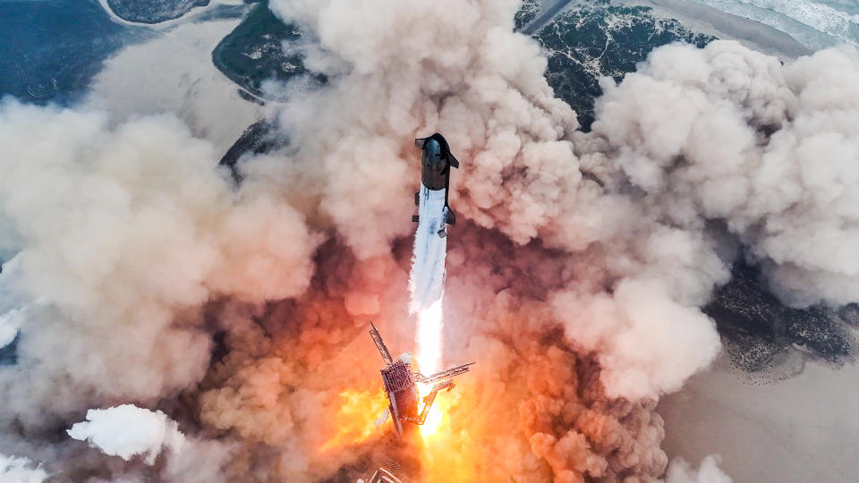  Drone's-eye view of a giant rocket launching from a seaside pad, generating a huge plume of dust and exhaust. 