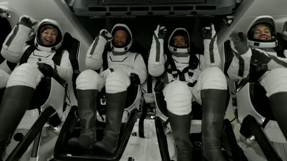  Four astronauts in white spacesuits smile and wave inside a cockpit. 