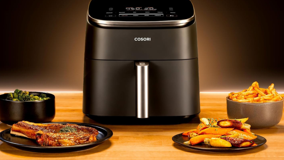 The Cosori air fryer on a table surrounded by assorted foods