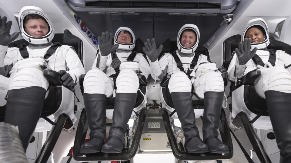 Four Crew-8 astronauts smile and wave in white SpaceX spacesuits inside their spacecraft.