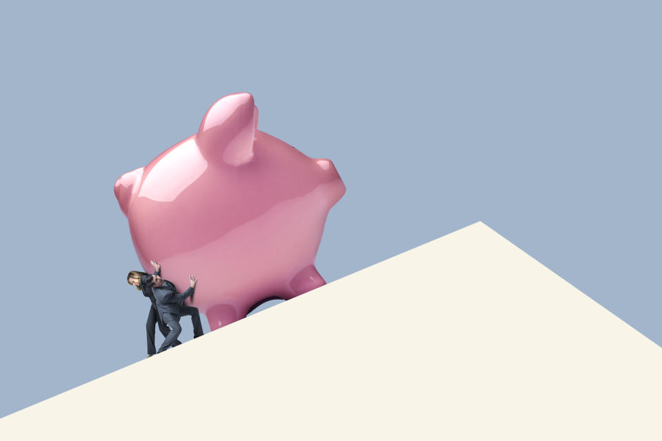 In a photo illustration, a man and a woman struggle as they attempt to push a piggy bank up a hill. 