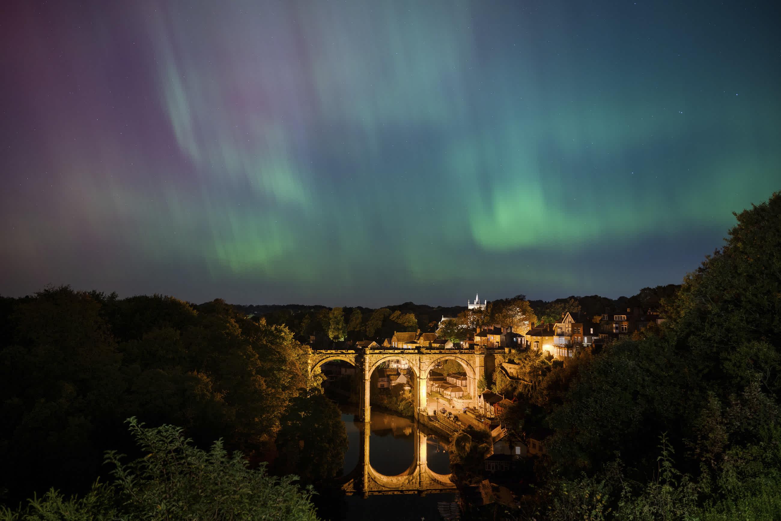 Aurora borealis against a tree-surrounded bridge.