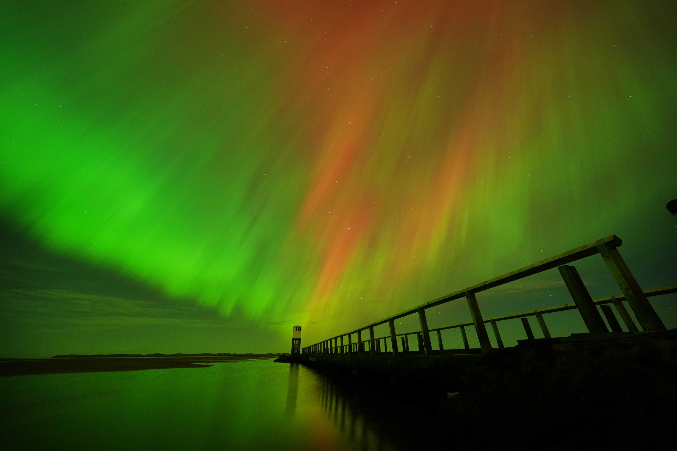 Aurora borealis against a silhouette of a walkway next to body of water.