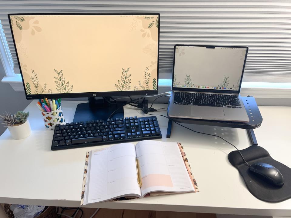 A desk with a laptop, external monitor, keyboard, mouse, pen organizer, a plant, and a planner on top.