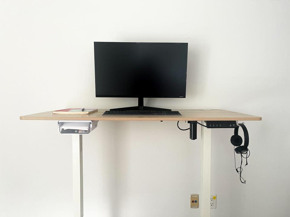 A computer monitor, keyboard, book, and pen on standing desk, with a pair of headphones hanging off the side.