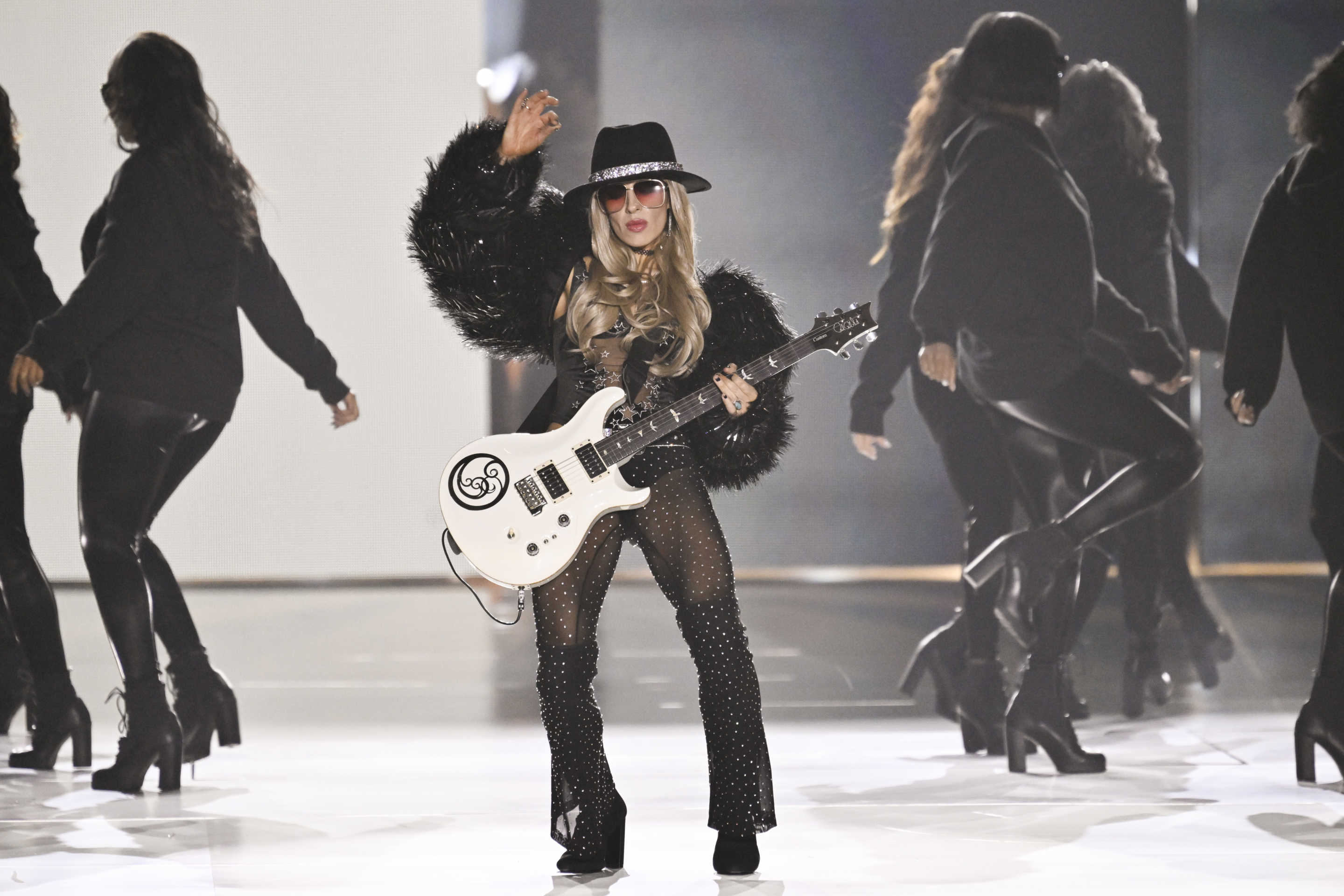 Orianthi, surrounded by models, performs in a black hat, black boots and black feathery jacket.