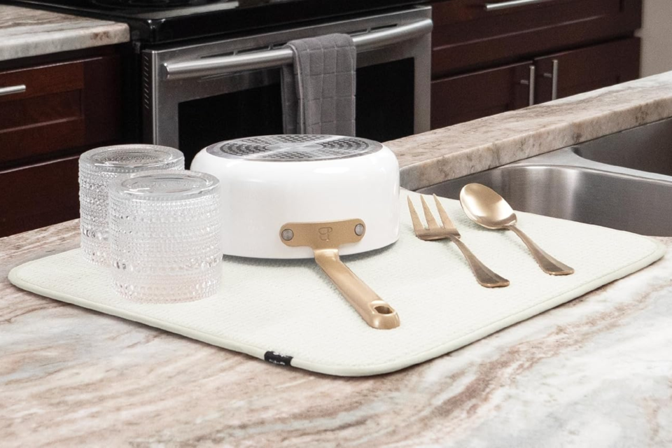 The cream-colored drying mat holding glasses, a pot and utensils on a kitchen counter