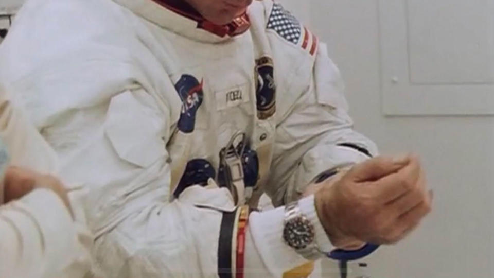 photo of an astronaut in a white spacesuit in a white-walled room on Earth. a watch is visible on the astronaut's wrist