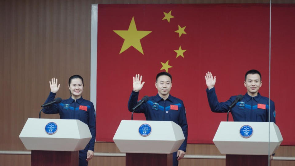 three astronauts in jumpsuits raise their right hands with bent elbows, standing behind white-capped podiums in front of a large chinese flag.