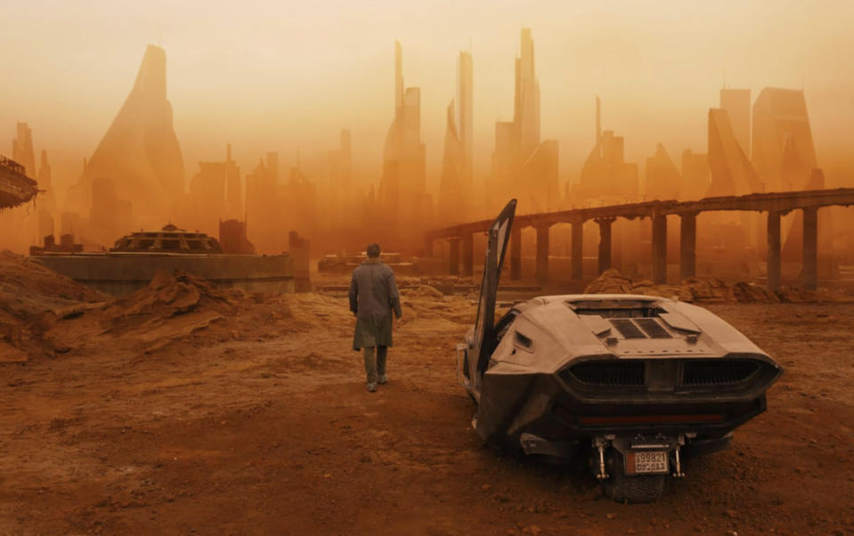 A man in a trench coat with a car and a desert landscape in the background.