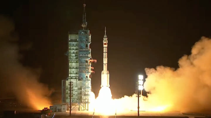  A white rocket launches next to a green tower at night, fire shooting from its bottom, a smoke plume blowing to the right. 