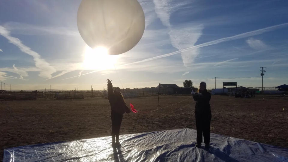  Two humans stand on a tarp on a grassy field as they release a large white balloon into the air, the sun glaring behind it as the contrails streak the blue sky above. 