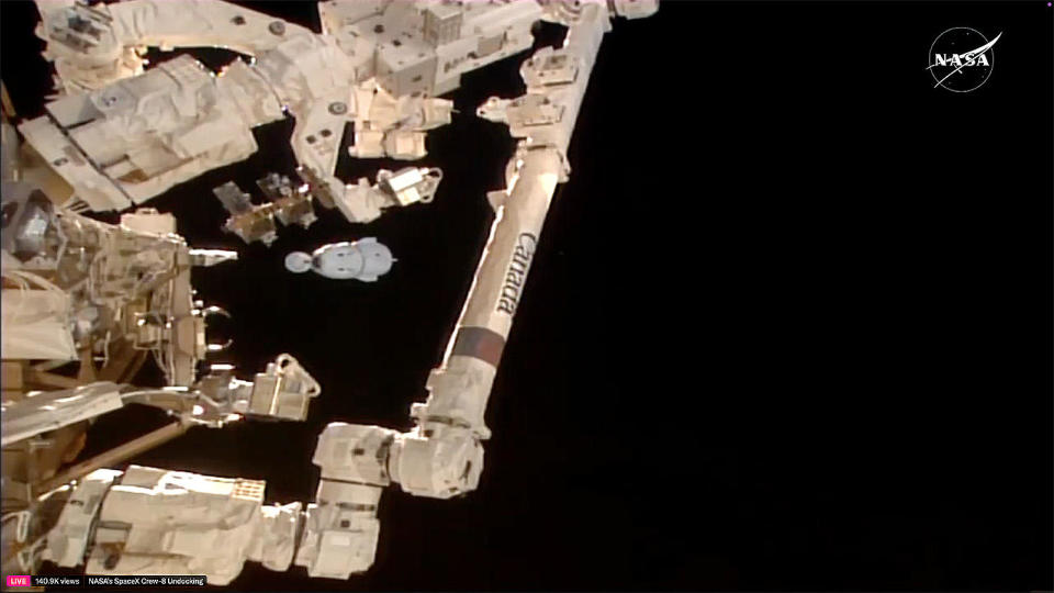 The SpaceX Crew Dragon ferry ship carrying three NASA astronauts and a Russian cosmonaut can be seen framed between segments of the International Space Station's robot arm moments after undocking from the International Space Station. Splashdown in the Gulf of Mexico is expected early Friday. / Credit: NASA TV