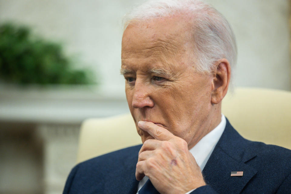 President Biden sits in an armchair in an official stateroom looking undecided, a finger to his pursed lips.