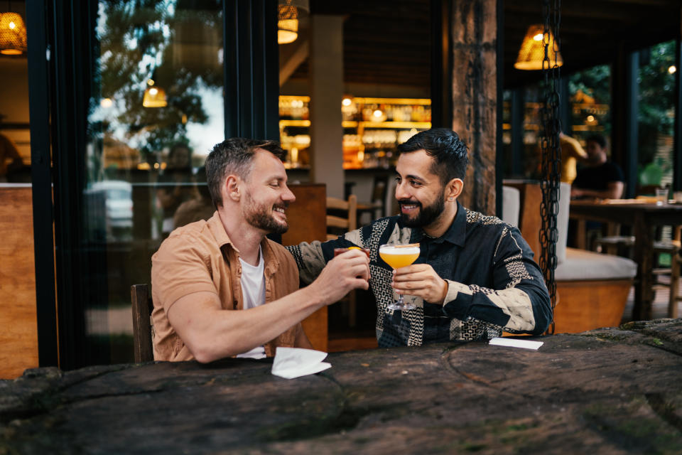 Young gay couple smiling while toasting with cocktails together outside at a table on a bar patio in the early evening