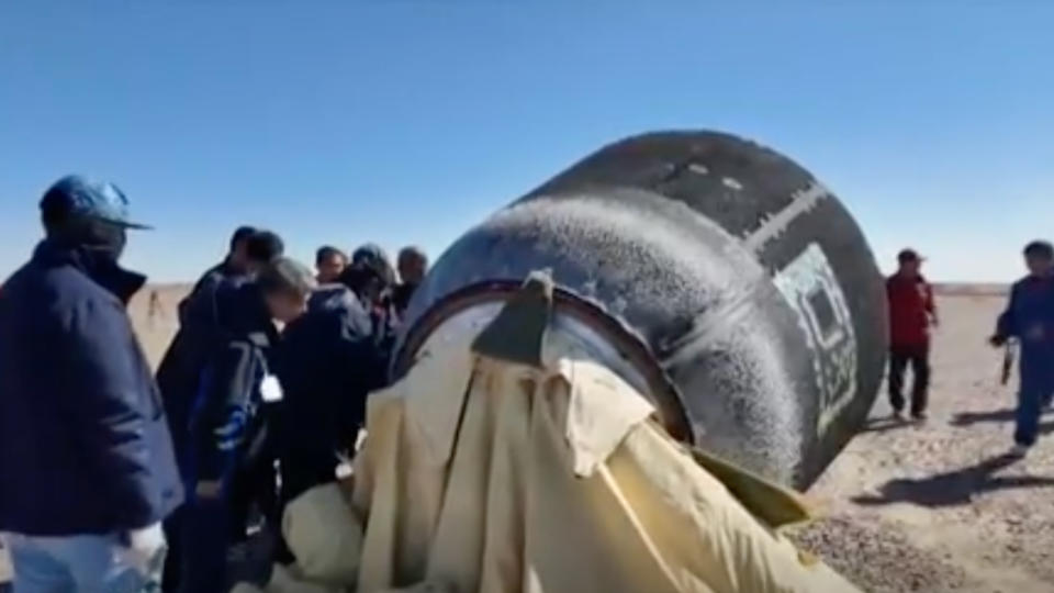  A group of people inspect a gray cylindrical spacecraft in a desert landscape. 