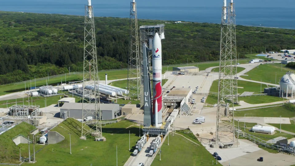  Aerial view of a red and white rocket on the launch pad, with greenery and the ocean in the background. 