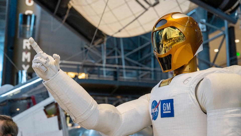  Closeup photo of a white humanoid robot with a gold helmet in a museum exhibit hall. 